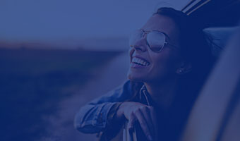 Frau mit Sonnenbrille schaut lächelnd aus dem Fenster eines fahrenden Autos
